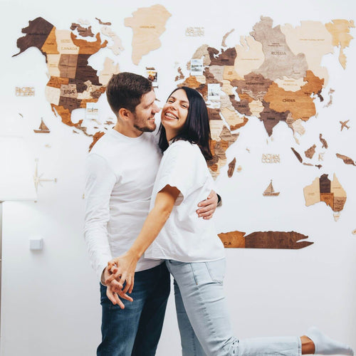 Happy couple embracing in front of a 3D wooden world map multicolor wall art with country names and small photos pinned